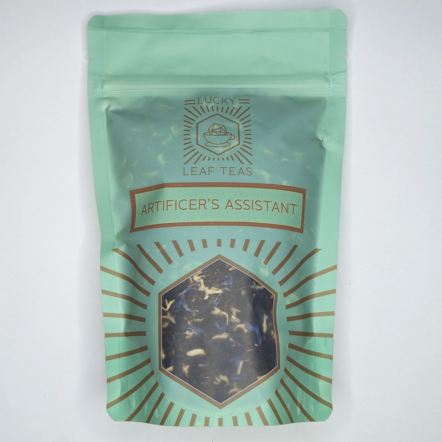 A mint-green package containing 100g of Artificer's Assistant: a black tea with visible yellow and blue cornflower petals. The name is stickered on the front, below the Lucky Leaf Teas Logo and above a hexagonal window displaying the tea.