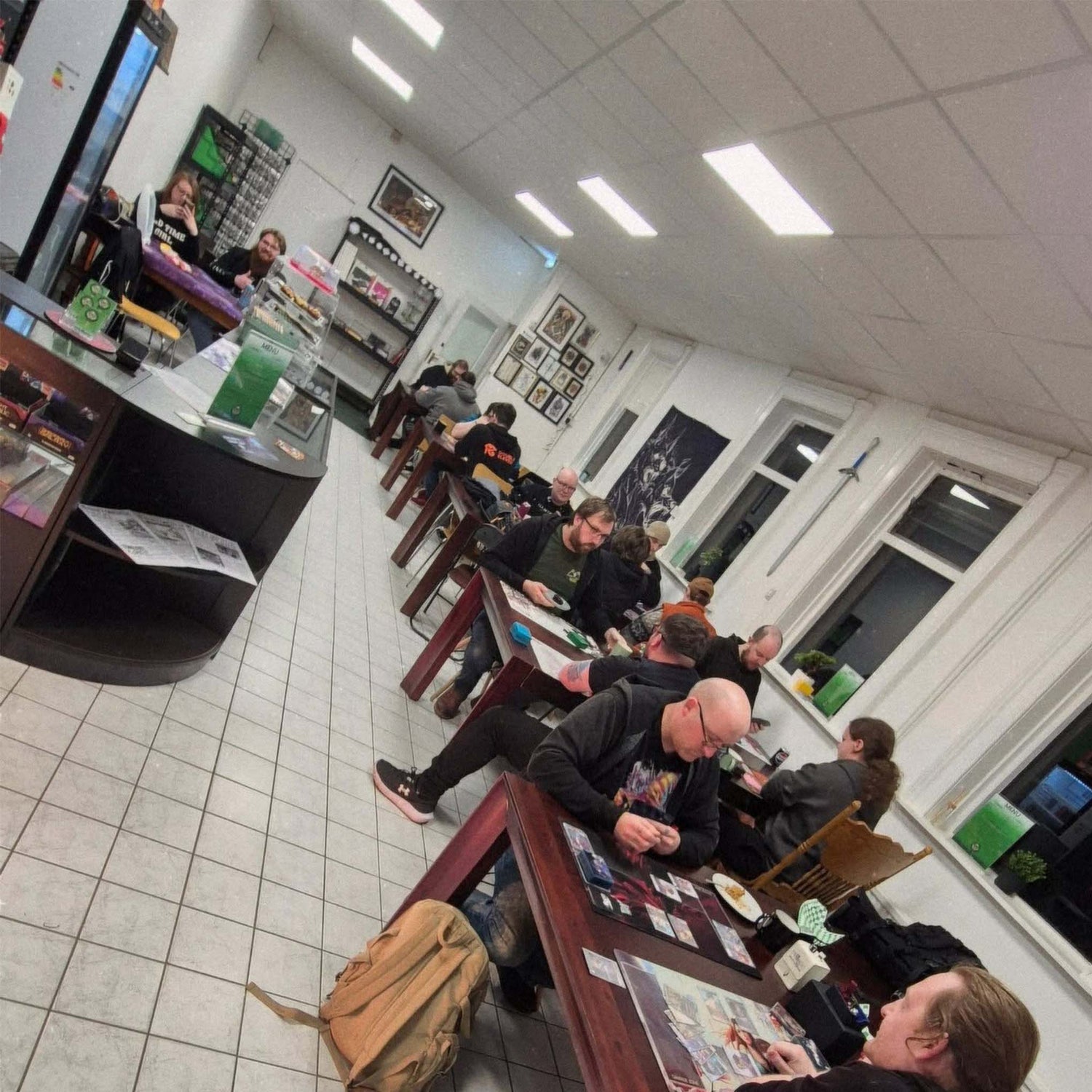 The inside of a boardgame cafe. On the right, several people sit at dark red wooden tables playing card games. On the left, a store counter, with a refrigerator and a pastry case.