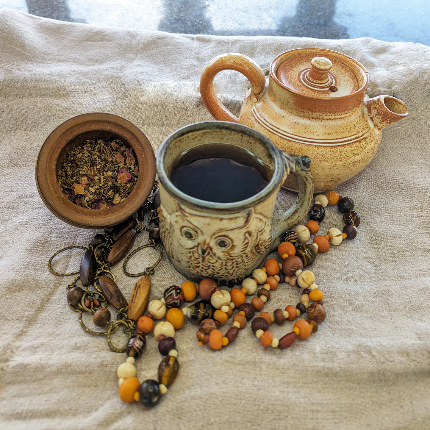 A clay teapot behind a rustic mug with an owl on it, filled with a darker brown tea. They are on a hessian sheet, alongside bead necklaces.
