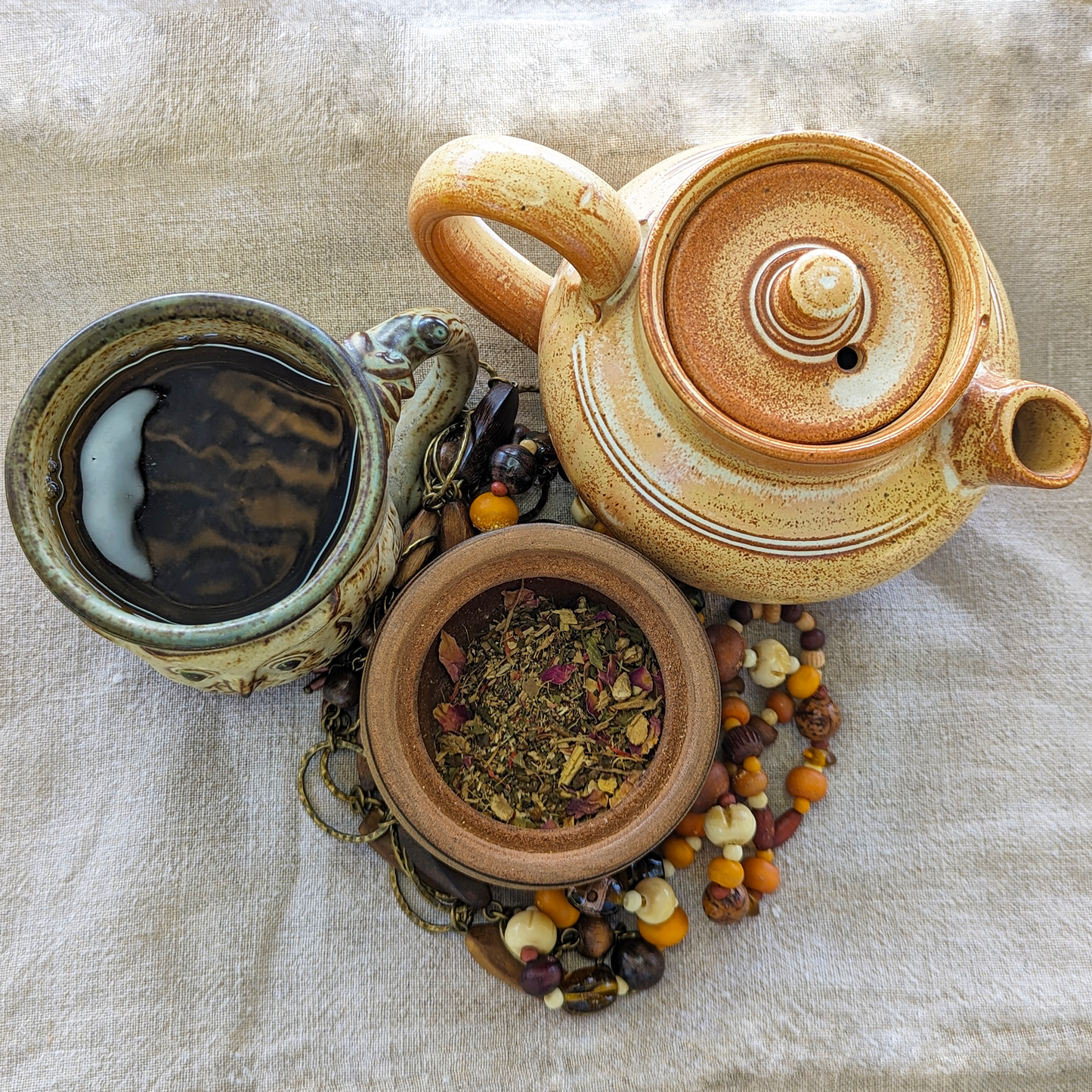 A clay teapot, rustic mug and wooden pot surround each other. The pot is filled with a herbal blend with rose petals and licorice root is visible in the pot.