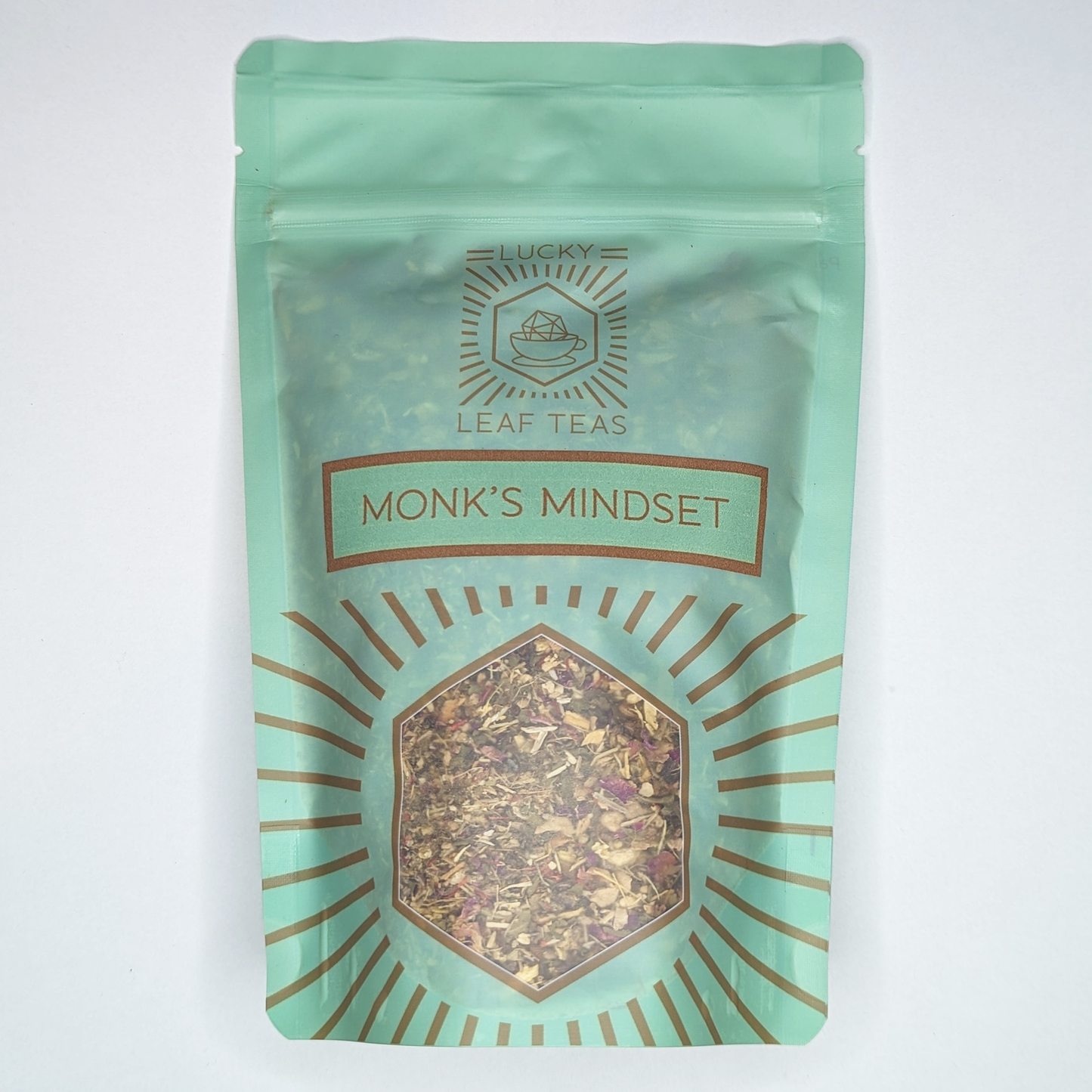 A mint-green package containing 100g of Monk’s Mindset: an infusion blend of herbs and flowers. The name is stickered on the front, below the Lucky Leaf Teas Logo and above a hexagonal window displaying the tea.