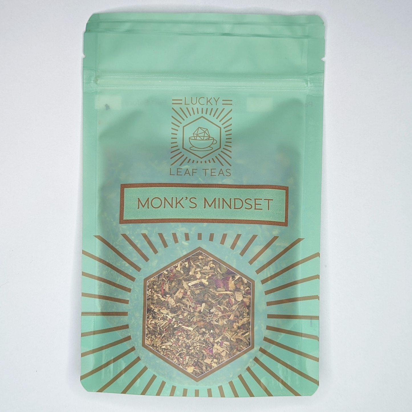 A mint-green package containing 50g of Monk’s Mindset: an infusion blend of herbs and flowers. The name is stickered on the front, below the Lucky Leaf Teas Logo and above a hexagonal window displaying the tea.