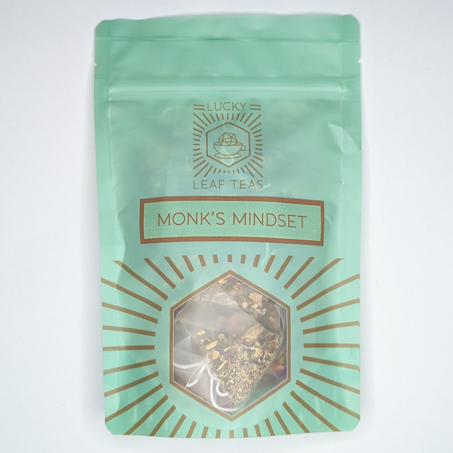A mint-green package containing 15 Silky Pyramid Teabags of Monk’s Mindset: an infusion blend of herbs and flowers. The name is stickered on the front, below the Lucky Leaf Teas Logo and above a hexagonal window displaying the tea.