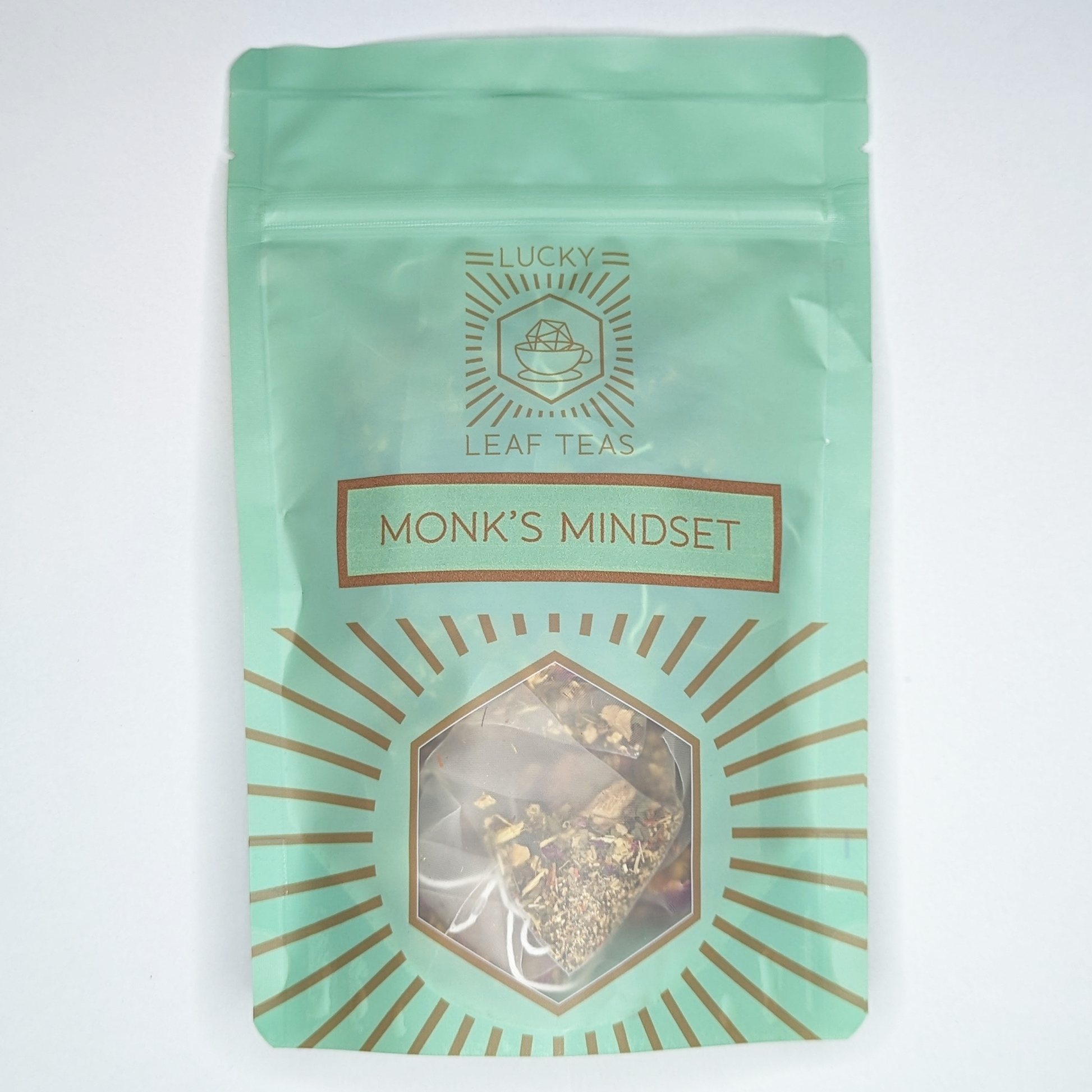 A mint-green package containing 15 Silky Pyramid Teabags of Monk’s Mindset: an infusion blend of herbs and flowers. The name is stickered on the front, below the Lucky Leaf Teas Logo and above a hexagonal window displaying the tea.
