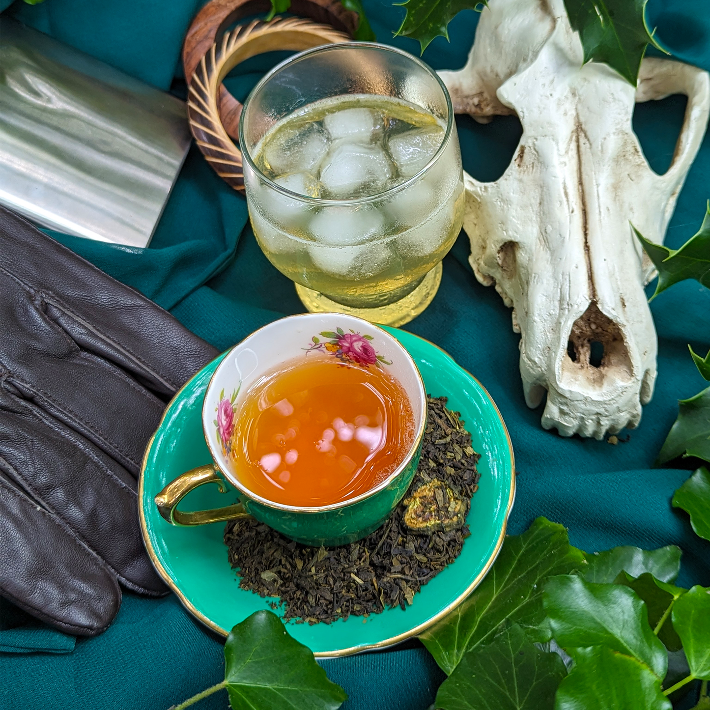 A cup of tea and a glass of ice tea, surrounded by plants, a hip flask, a pair of leather gloves and a fox's skull.