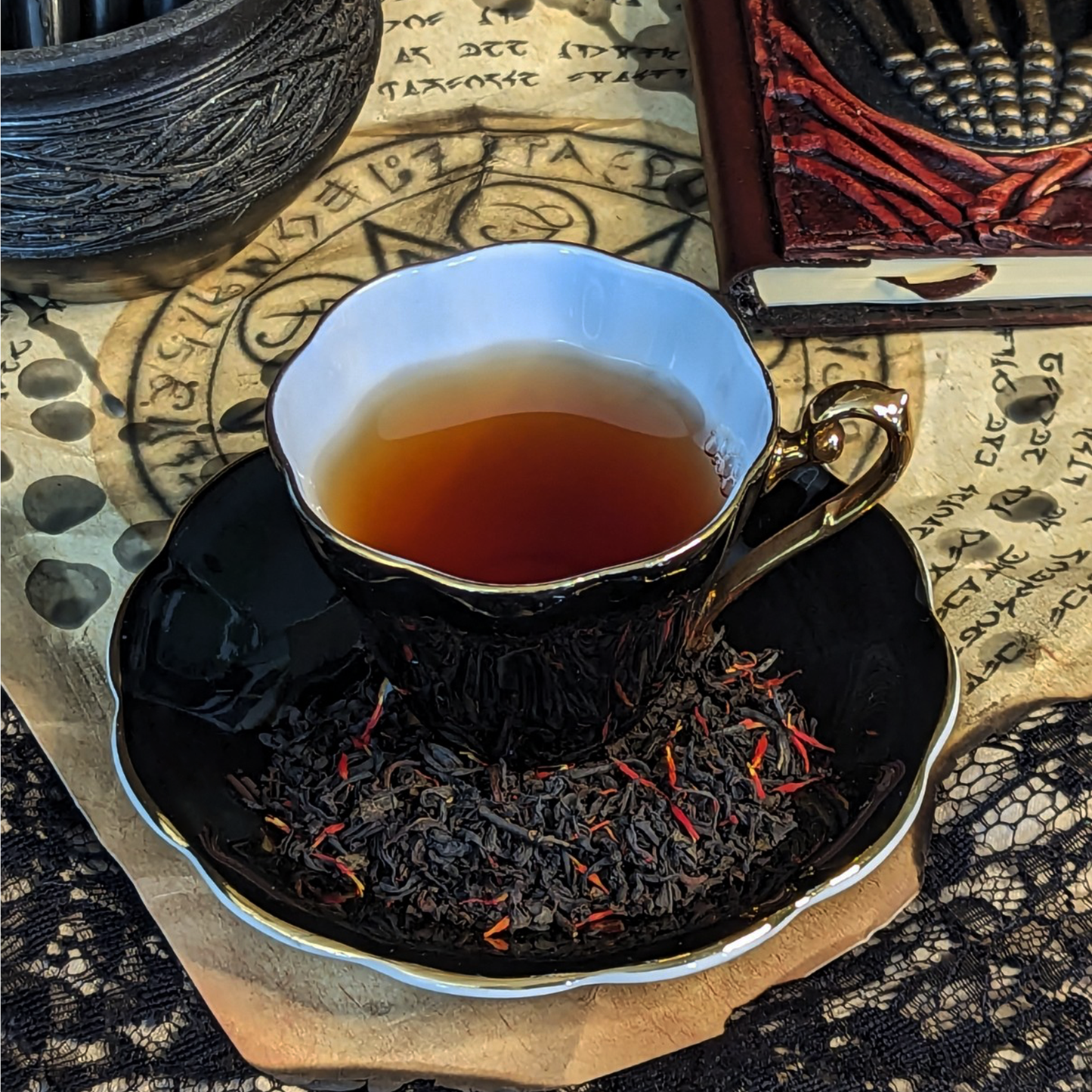 A black teacup of tea and a matching saucer on top of wax-covered aged paper with summoning circles on it. Tea leaves mixed with red flowers are on the saucer.