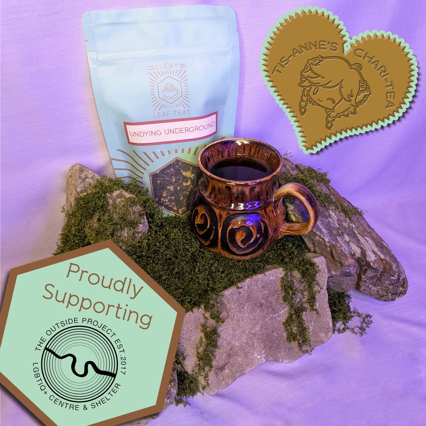 A brown earthenware mug with a dark brown liquid inside sits on top of a bed of moss and lichen-covered stones. Behind it stands a mint-green pouch with the Lucky Leaf Teas logo, and the name "Undying Underground" on the front. The loose leaf blend can be seen within. A heart-shaped badge in one corner reads "Tis-Anne's Chari-Tea", and another emblem reads "Proudly Supporting" with the logo for The Outside Project.