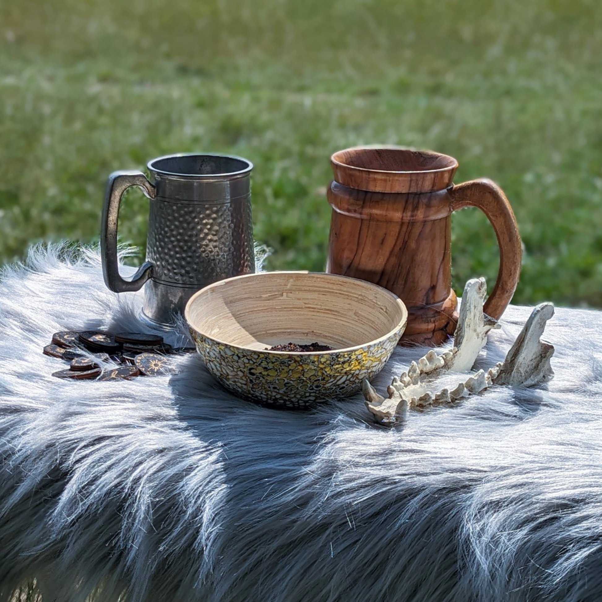 Two flagons, a wooden bowl, a handful of black wooden runes and a sheep's skull jawbone are on top of a grey fleece. Grass is in the background.