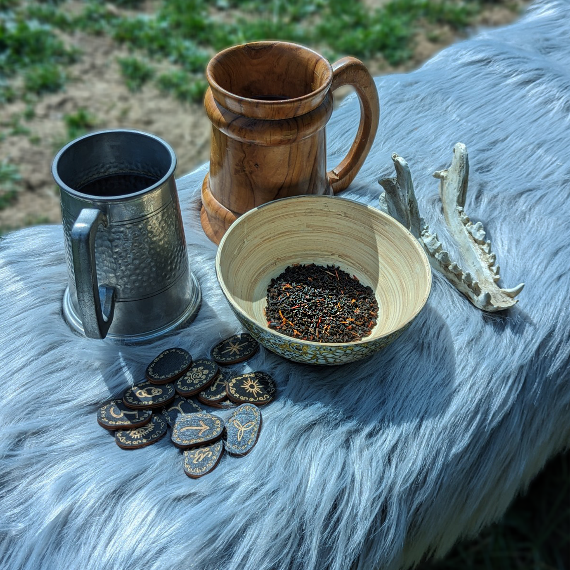 Two flagons, a wooden bowl of tea leaves, a handful of black wooden runes and a sheep's skull jawbone on top of a grey fleece.