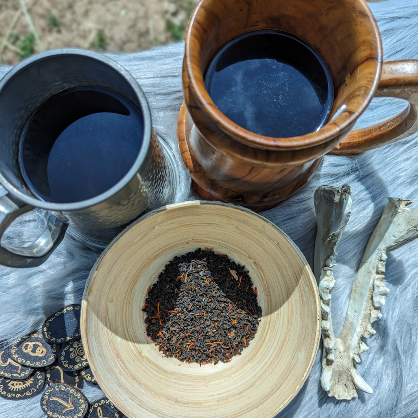 A close-up of two flagons, a wooden bowl of tea leaves, a handful of black wooden runes and a sheep's skull jawbone on top of a grey fleece. The flagons are filled with dark tea.