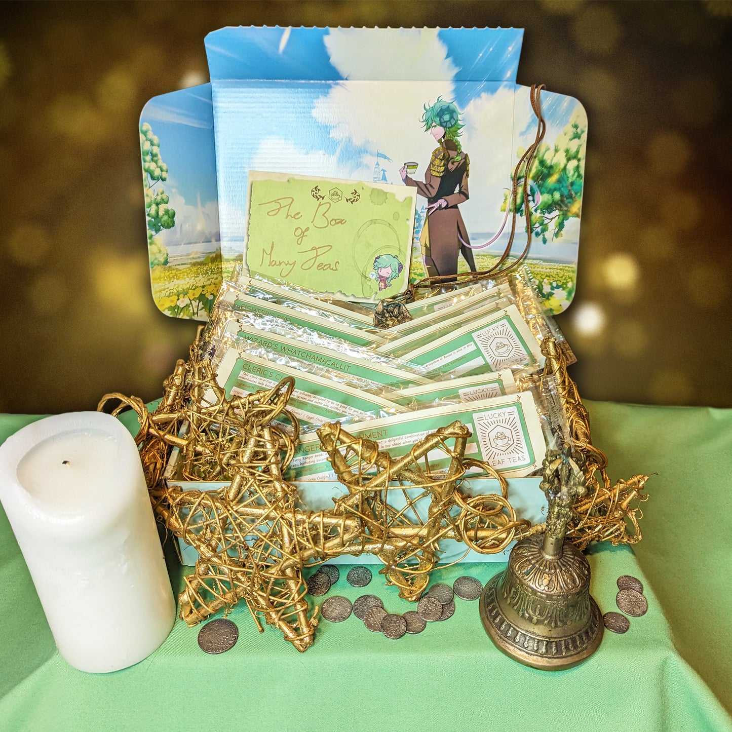 An open mint-coloured box, with an illustration of a horned character with a teacup smiling towards the viewer. The box is filled with slim packages of tea and herbal blends, and is surrounded by a small brass bell, an unlit wax candle, small silver coins, and a wreath of golden wire stars. A card on the box reads: "The Box of Many Teas."