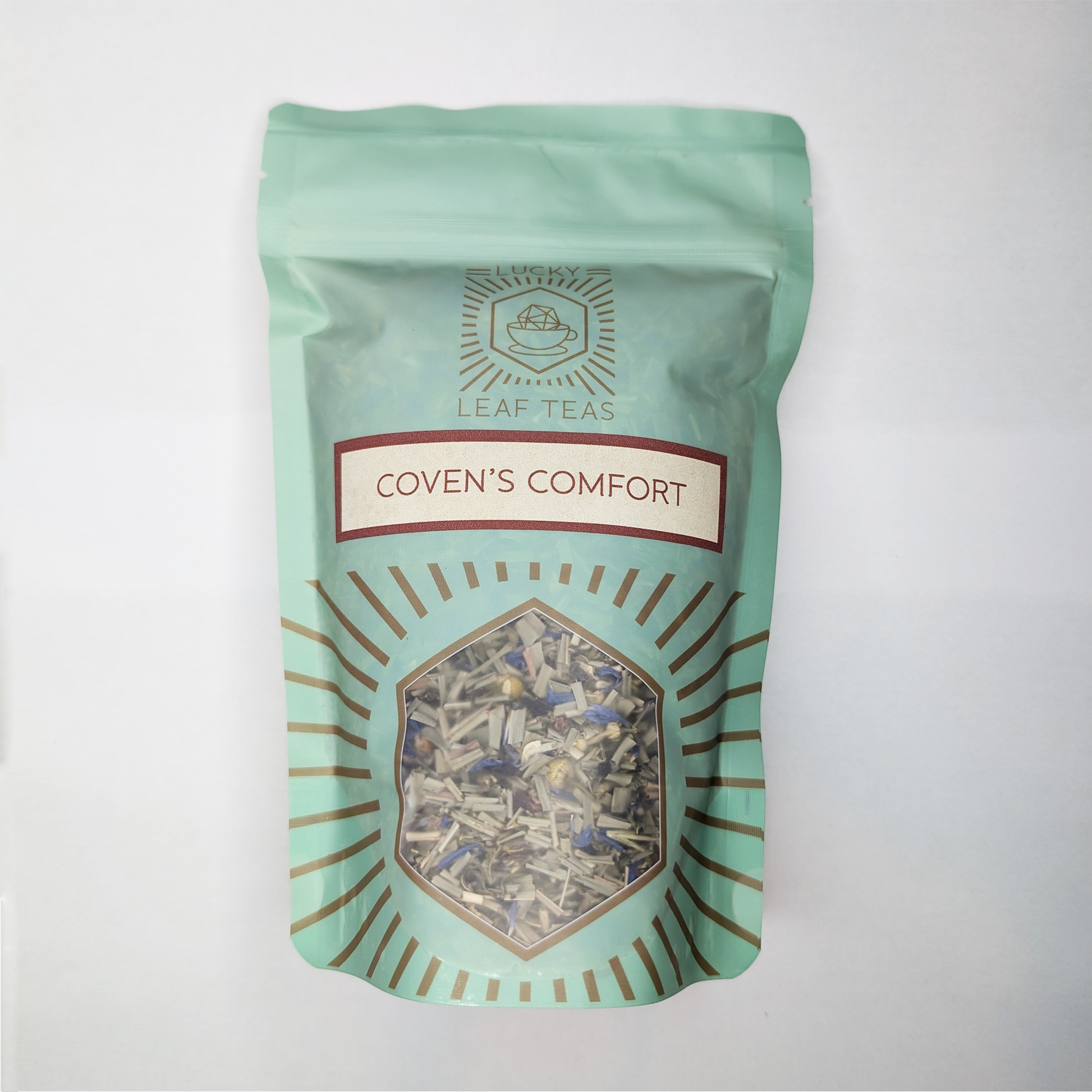 A mint-green package containing 100g of Coven's Comfort: a herbal infusion. The name is stickered on the front, below the Lucky Leaf Teas Logo and above a hexagonal window displaying the tea.