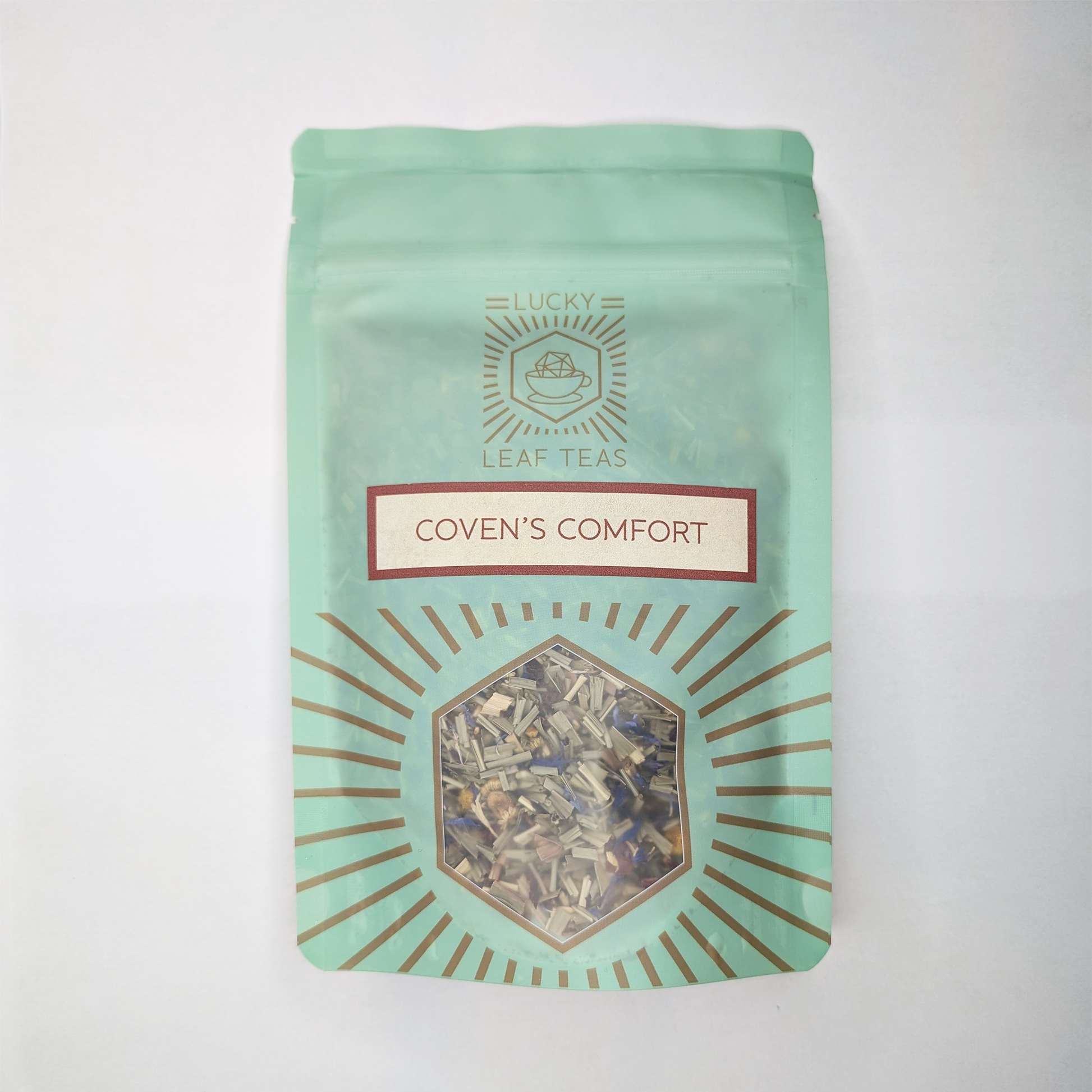 A mint-green package containing 50g of Coven's Comfort: a herbal infusion. The name is stickered on the front, below the Lucky Leaf Teas Logo and above a hexagonal window displaying the tea.