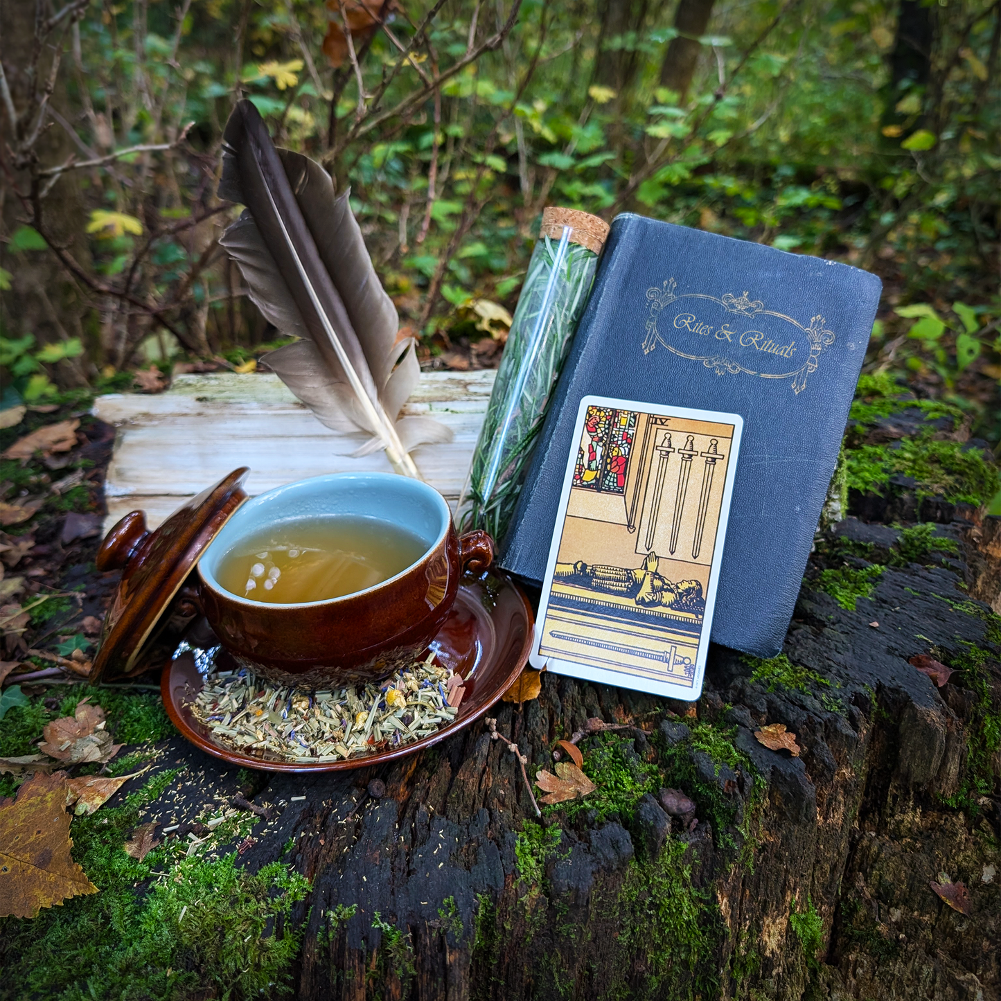 A brown-glazed bowl filled with a clear, amber-yellow liquid. It sits on a matching saucer, which has a scattering of leaves and flowers upon it. To the right is a "Four of Swords" tarot card, a pair of feathers, a corked vial filled with a fresh herb, and a blue book titled "Rites and Rituals."