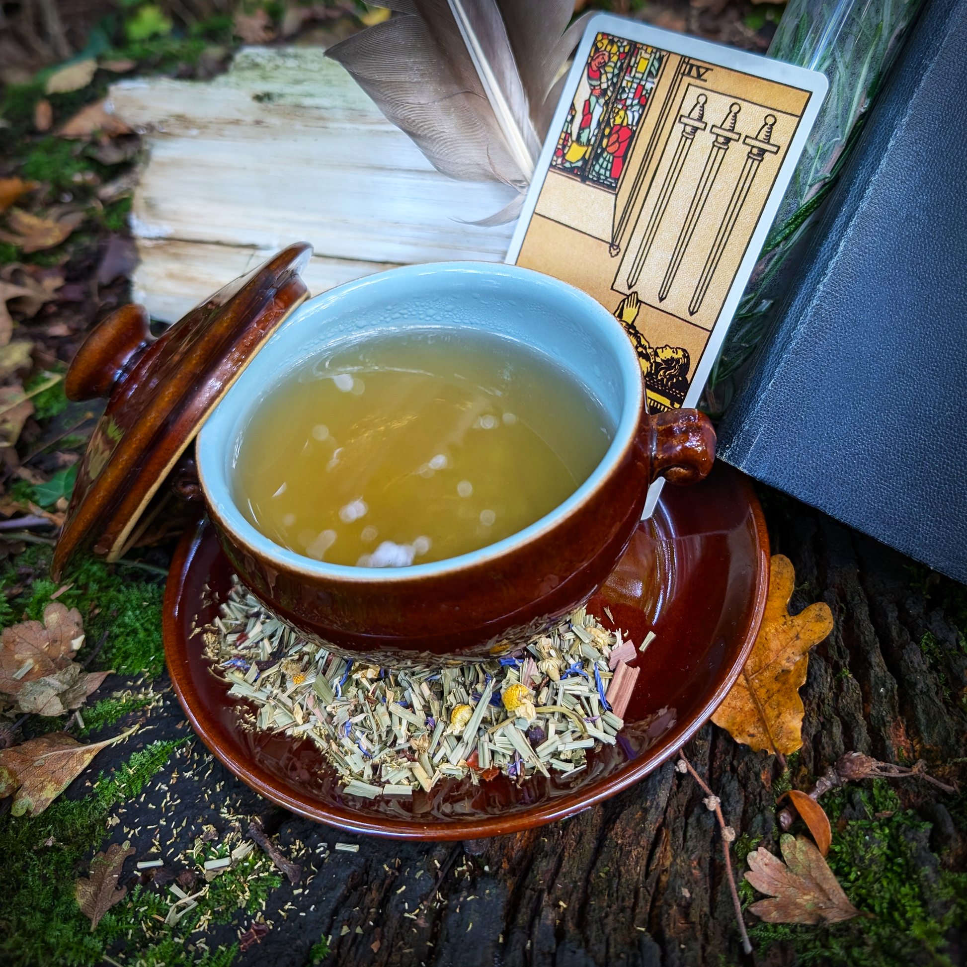 A brown-glazed bowl filled with a clear, amber-yellow liquid. It sits on a matching saucer, which has a scattering of leaves and flowers upon it. They, as well as a "Four of Swords" tarot card, sit on top of a mossy log.