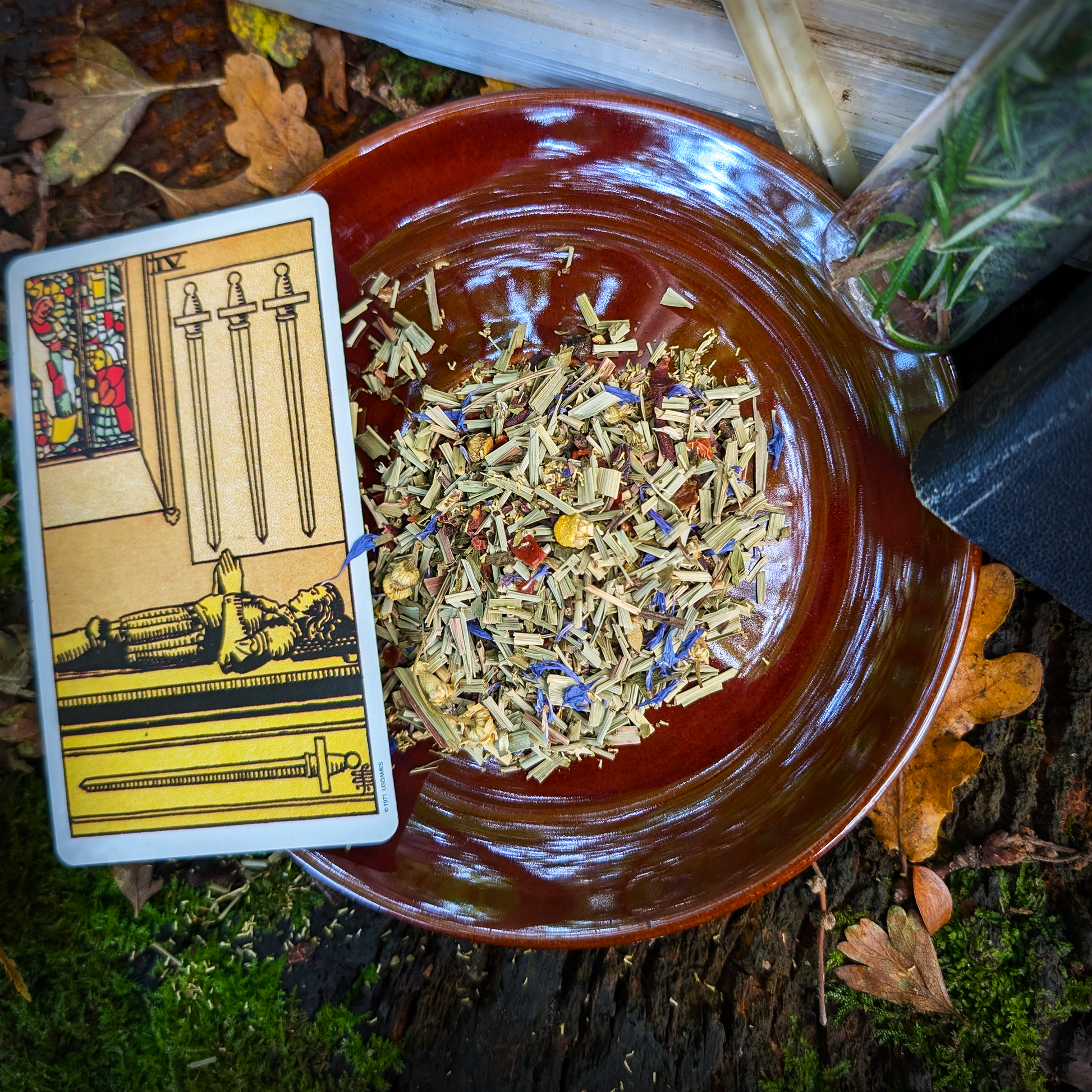 A brown-glazed saucer with a mound of leaves and flowers, a herbal infusion, on top of it. Delicately to the side is a "Four of Swords" tarot card.