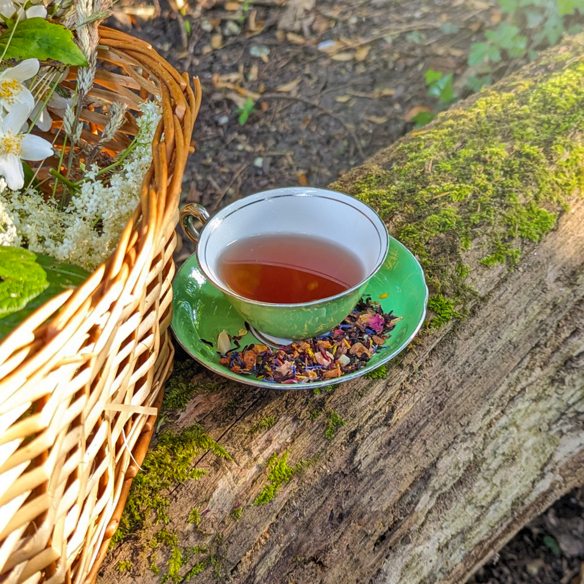 A green teacup of amber tea and a matching saucer on a moss-covered log. A basket of wildflowers is next to it.