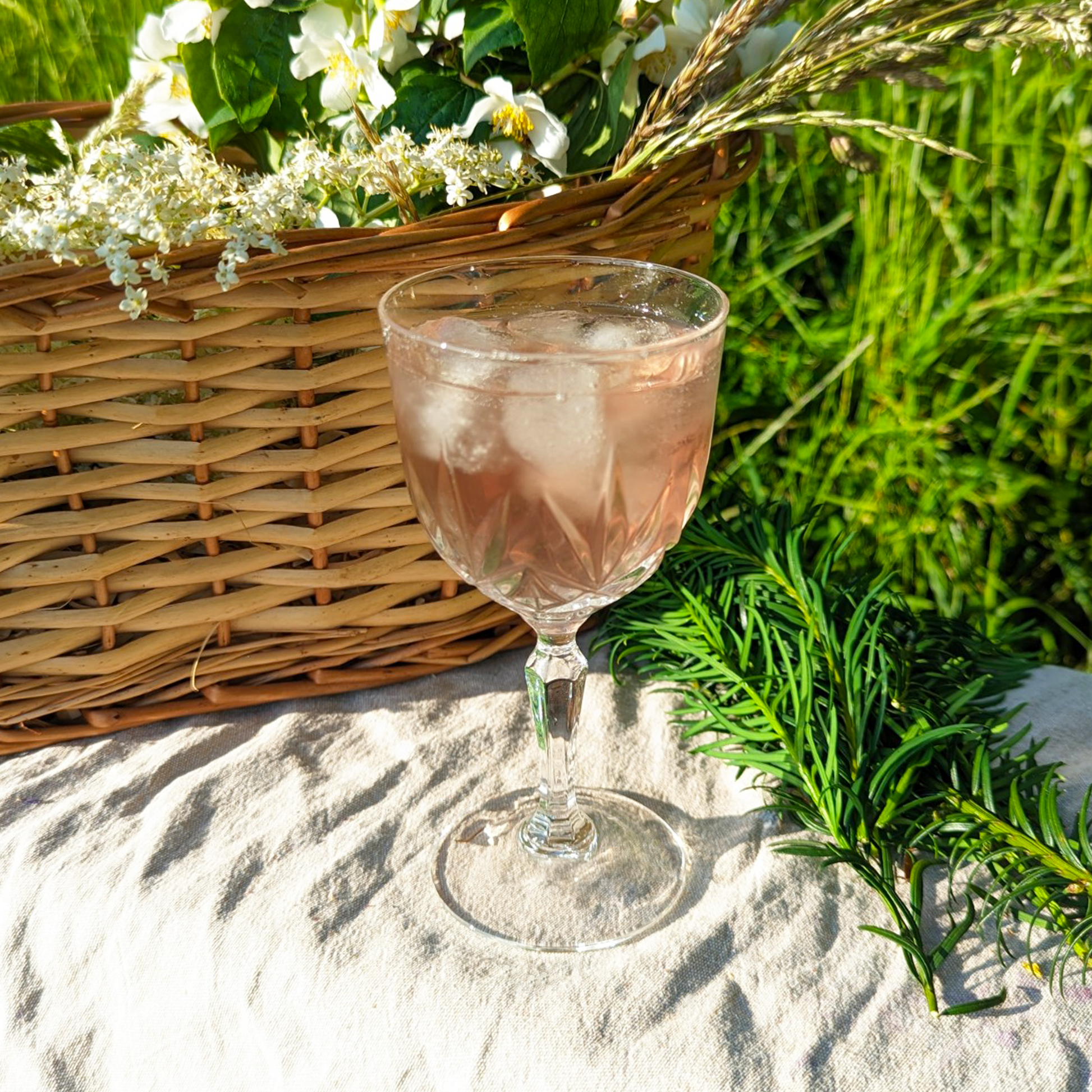 Peach-coloured tea in a glass, with ice. A basket of wildflowers is behind it, with lots of grass nearby.