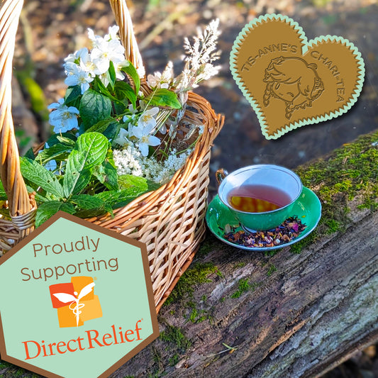 A green teacup and saucer on  a mossy log, next to a basket of wildflowers and leaves. In the top right is a heart-shaped badge reading "Tis-Anne's Chari-Tea" and in the bottom left a hexagonal badge reading "Proudly Supporting Direct Relief."