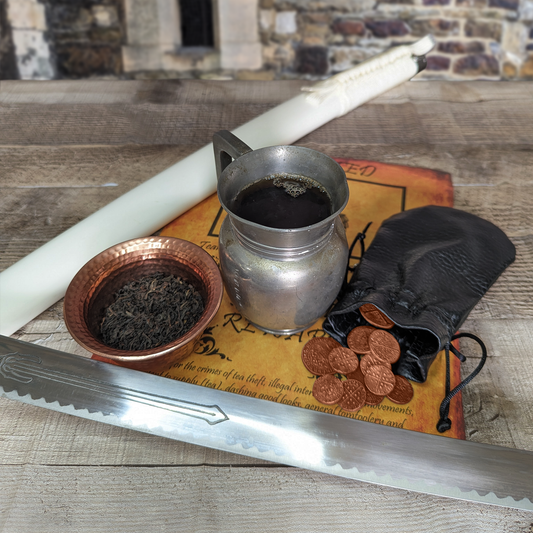 An old tin mug filled with dark tea, next to a pouch of copper coins and a copper dish filled with black tea leaves. The blade of a sword and its sheath surround these items.