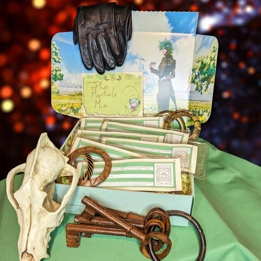 An open mint-coloured box, with an illustration of a horned character with a teacup smiling towards the viewer. The box is filled with slim packages of tea and herbal blends, and is surrounded by a rusty set of keys, a fake fox's skull, a pair of leather gloves and several carved-wood and metal bracelets. A card on the box reads: "The Martial's Mix."