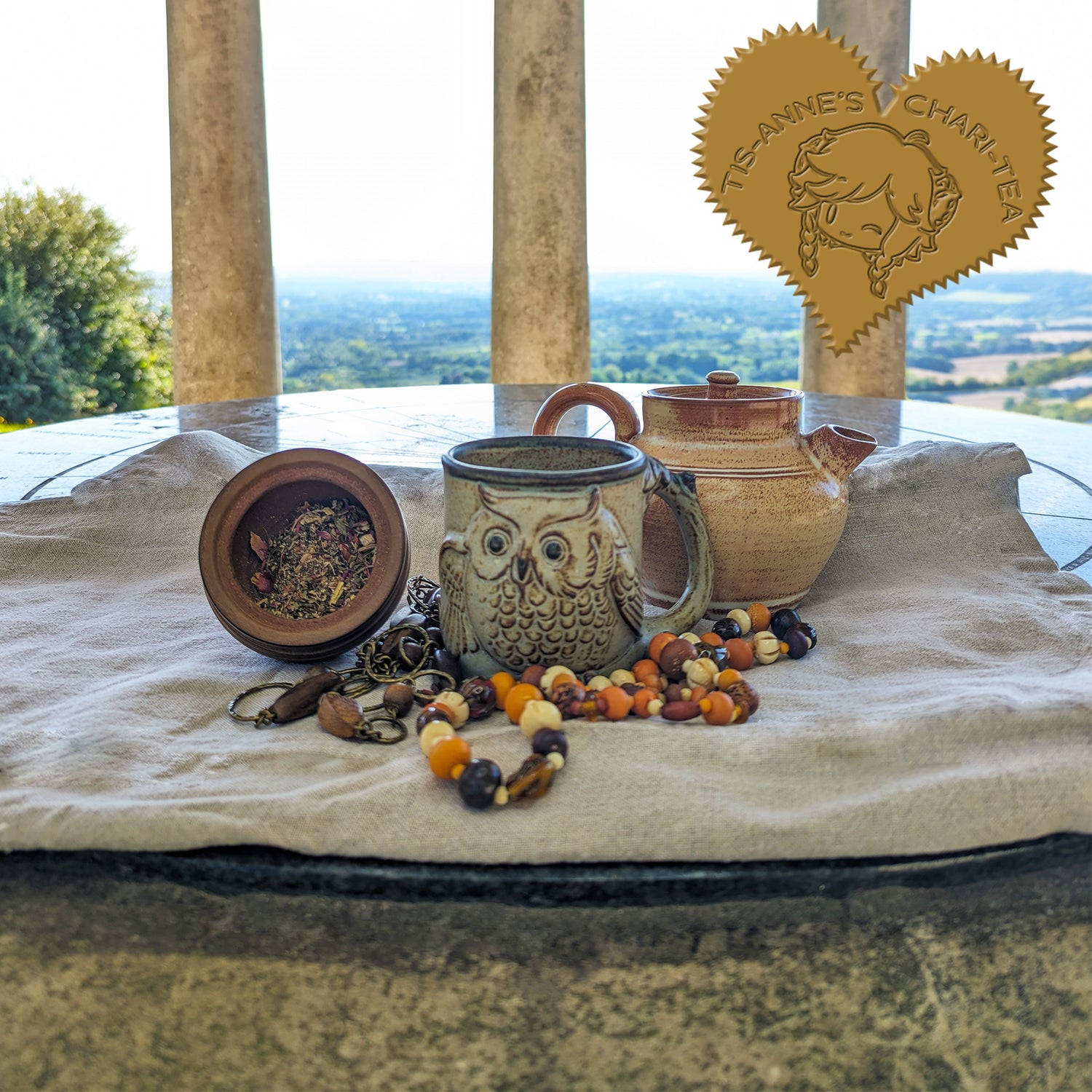 A clay teapot behind a rustic mug with an owl on it. Next to it is a wooden pot filled with a herbal blend. Stone pillars are in the background, with fields and trees in the distance. A heart-shaped badge in the top left corner reads "Tis-Anne's Chari-Tea".
