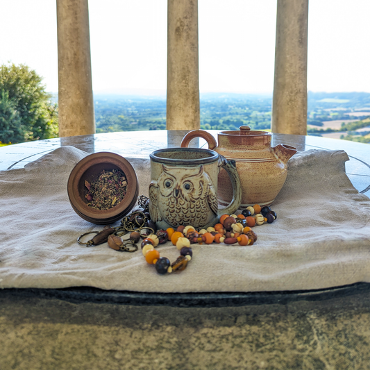 A clay teapot behind a rustic mug with an owl on it. Next to it is a wooden pot filled with a herbal blend. Stone pillars are in the background, with fields and trees in the distance.