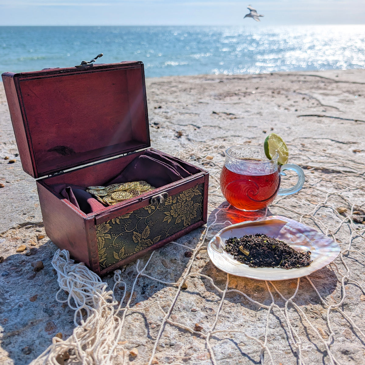 An open red-wood box with a decorative front reveals gold coins on a red satin pillow. Adjacent is a mother-of-pearl shell filled with tea leaves and behind sits a glass cup with a wave motif, filled with amber-red tea and a lime wedge on the rim. In the background you can see the sea and sky, and a bird flying past. Everything sits on a rope net.