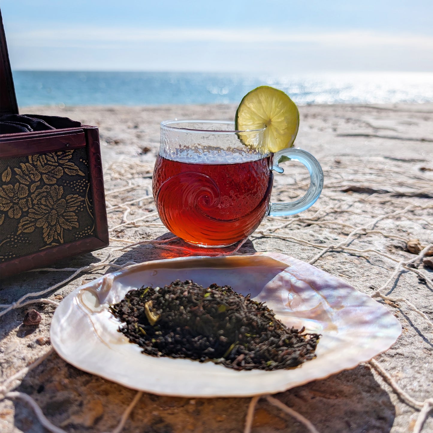 Atop a rope net sits a few objects. In the foreground is a mother-of-pearl shell filled with tea leaves and behind sits a glass cup with a wave motif, filled with amber-red tea and a lime wedge on the rim. Distantly, you can see the ocean and sky.