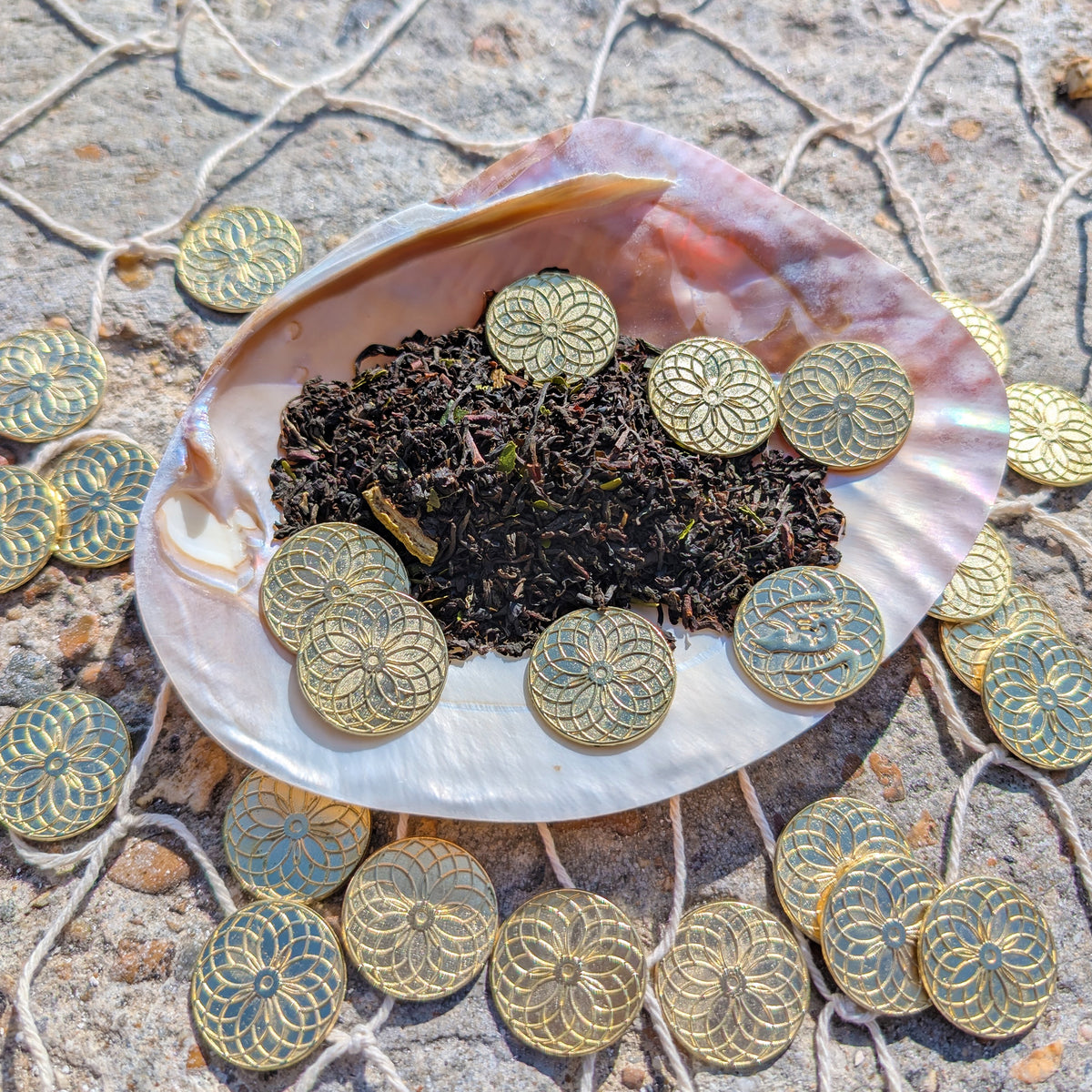 A mother-of-pearl shell sits on top of rope netting on a rocky surface. The shell is filled with small golden coins with a mandala design, as well as a small pile of black tea leaves flecked with green and lime rinds.