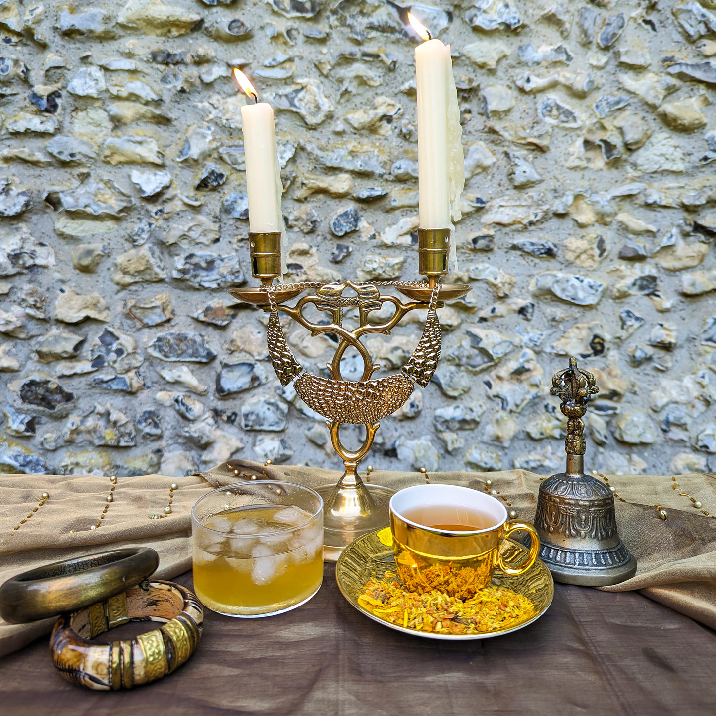 A golden cup of tea sits on a matching saucer, next to a glass cup of iced tea. A brass candlestick burns behind them.