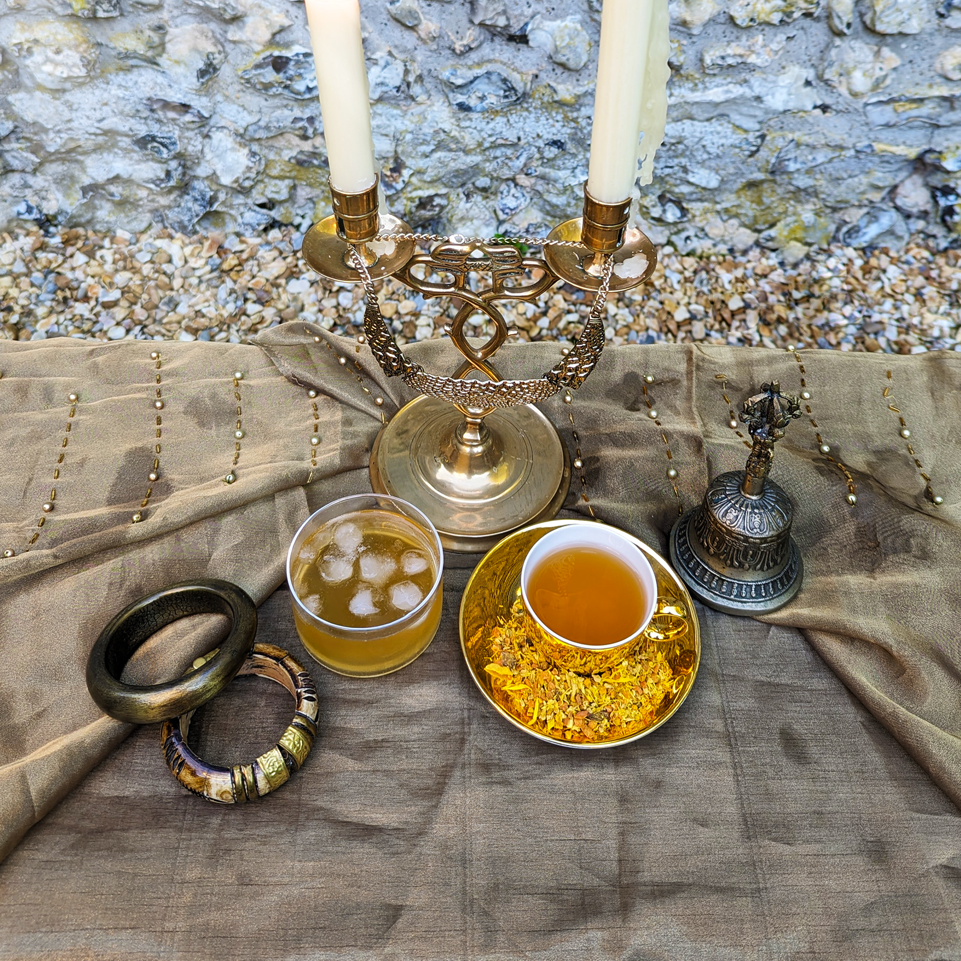 A golden cup of tea sits on a matching saucer, next to a glass cup of iced tea, on top of a gold satin sheet.