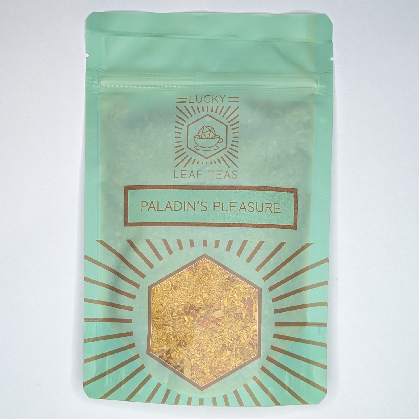 A mint-green package containing 100g of Paladin’s Pleasure: a bright yellow infusion blend of spices, fruits and flowers. The name is stickered on the front, below the Lucky Leaf Teas Logo and above a hexagonal window displaying the tea.