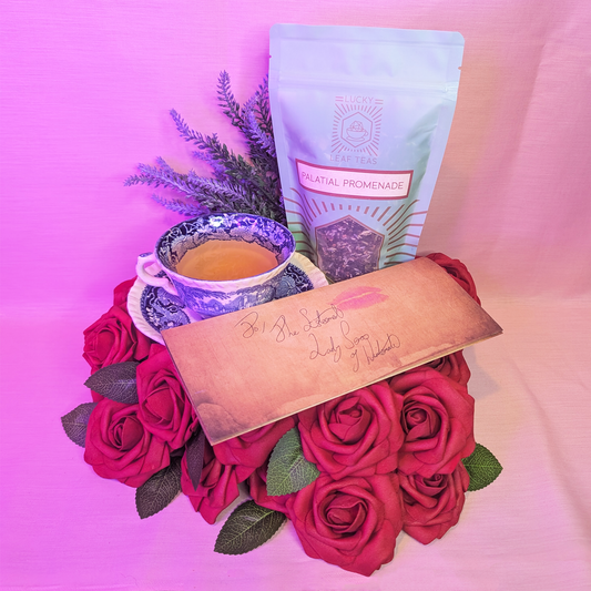 A blue china teacup filled with a pale yellow liquor is flanked by a folded letter addressed to "The Esteemed Lady Seros of Wintermade". The letter has a lipstick mark kiss. They sit on a bed of roses, alongside a mint-green pouch with the Lucky Leaf Teas logo, and the name "Palatial Promenade" on the front. The loose leaf blend can be seen within. Behind, sprigs of lavender poke out.