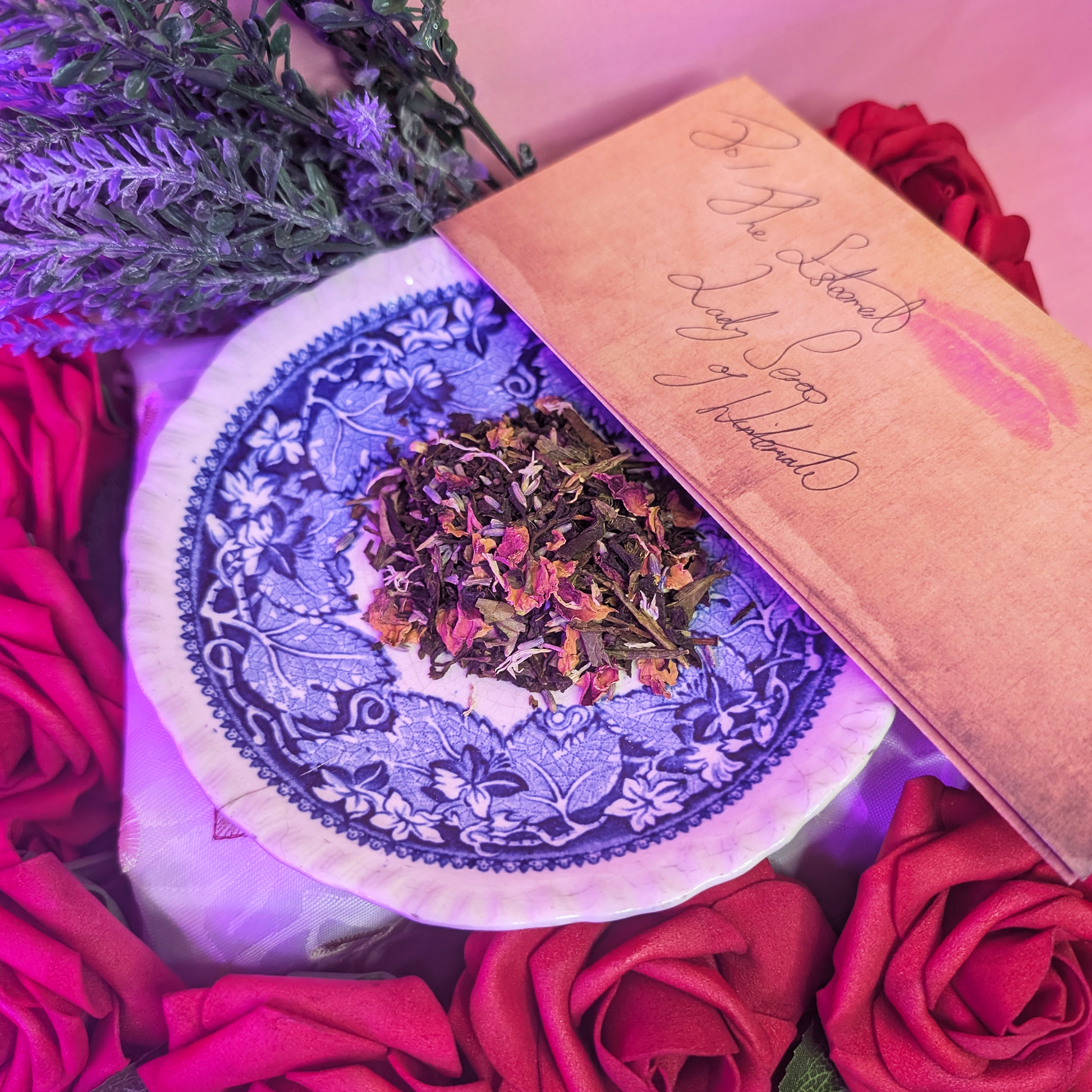 A blue china saucer with a mound of tea leaves and flower petals atop it. A folded letter addressed to "The Esteemed Lady Seros of Wintermade" with a lipstick mark is adjacent. They sit on a bed of roses with sprigs of lavender behind.