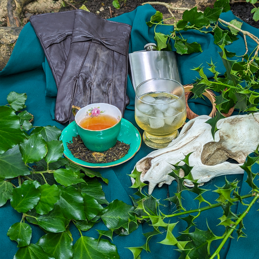 A cup of tea and a glass of ice tea, surrounded by plants, a hip flask, a pair of leather gloves and a fake fox's skull.