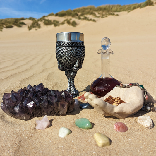 A goblet covered in silver dragon scales next to a tall-necked glass bottle filled with a dark red infusion. They are half-buried in the sand, with a blue sky above.