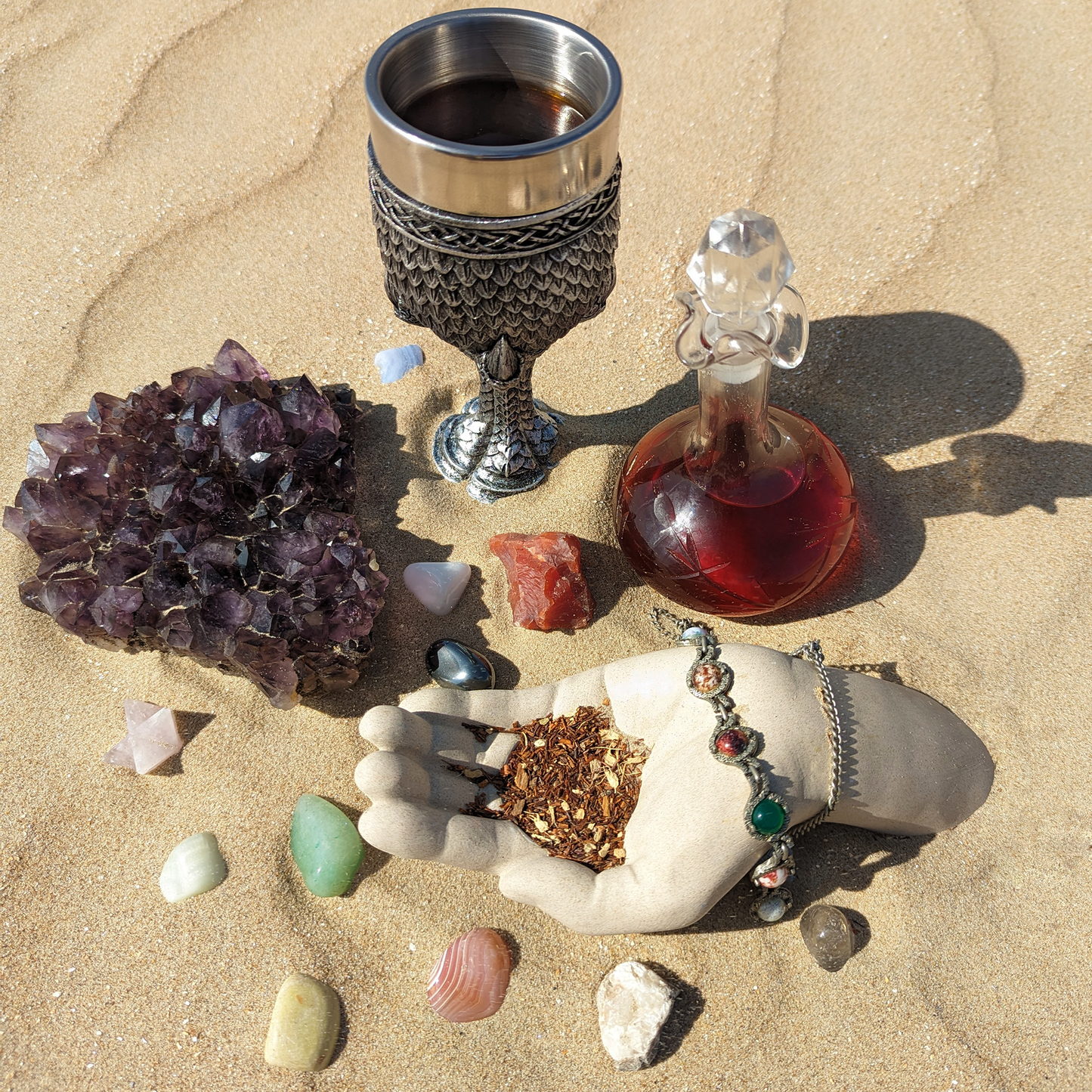 A goblet and glass bottle filled with dark-red and ruby-red liquids. They are surrounded by precious stones, a chunk of amethyst and a cement hand cupping a herbal blend.