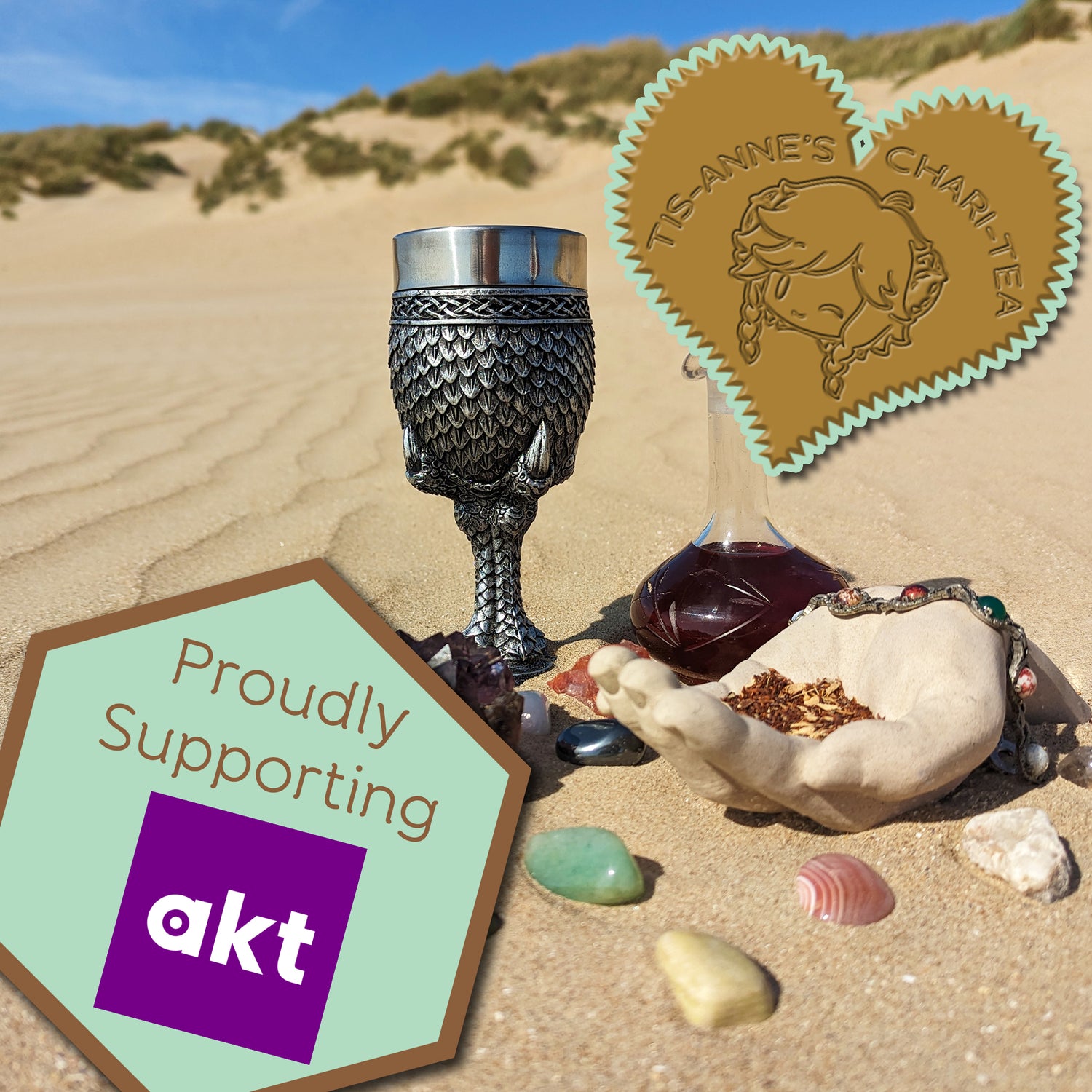 A goblet covered in silver dragon scales next to a tall-necked glass bottle filled with a dark red infusion. They are half-buried in the sand, with a blue sky above.
A heart-shaped badge in one corner reads "Tis-Anne's Chari-Tea", and an emblem in the other reads "Proudly Supporting AKT."