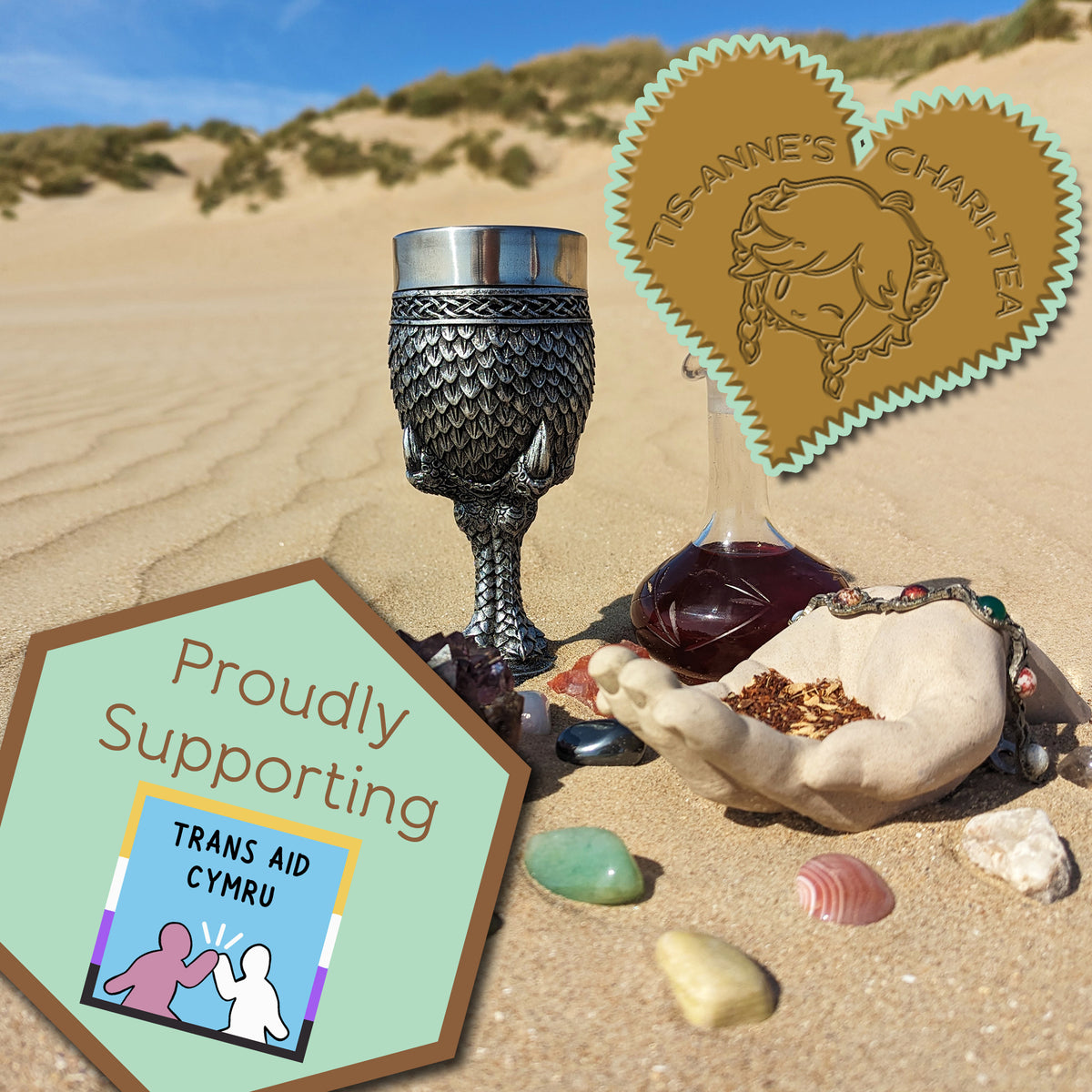 A goblet covered in silver dragon scales next to a tall-necked glass bottle filled with a dark red infusion. They are half-buried in the sand, with a blue sky above.
A heart-shaped badge in one corner reads "Tis-Anne's Chari-Tea", and an emblem in the other reads "Proudly Supporting - Trans Aid Cymru."