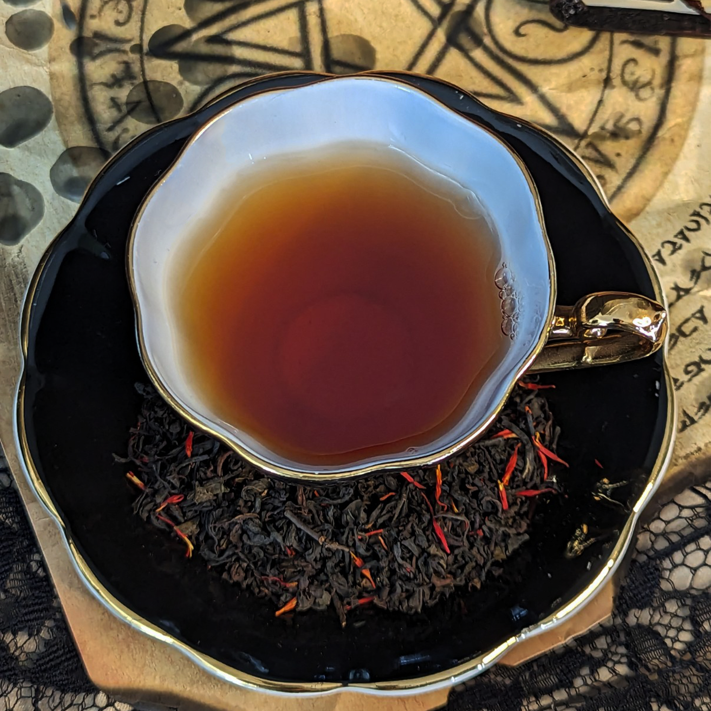 A black teacup of tea and a matching saucer on top of wax-covered aged paper with summoning circles on it. Tea leaves mixed with red flowers are on the saucer.