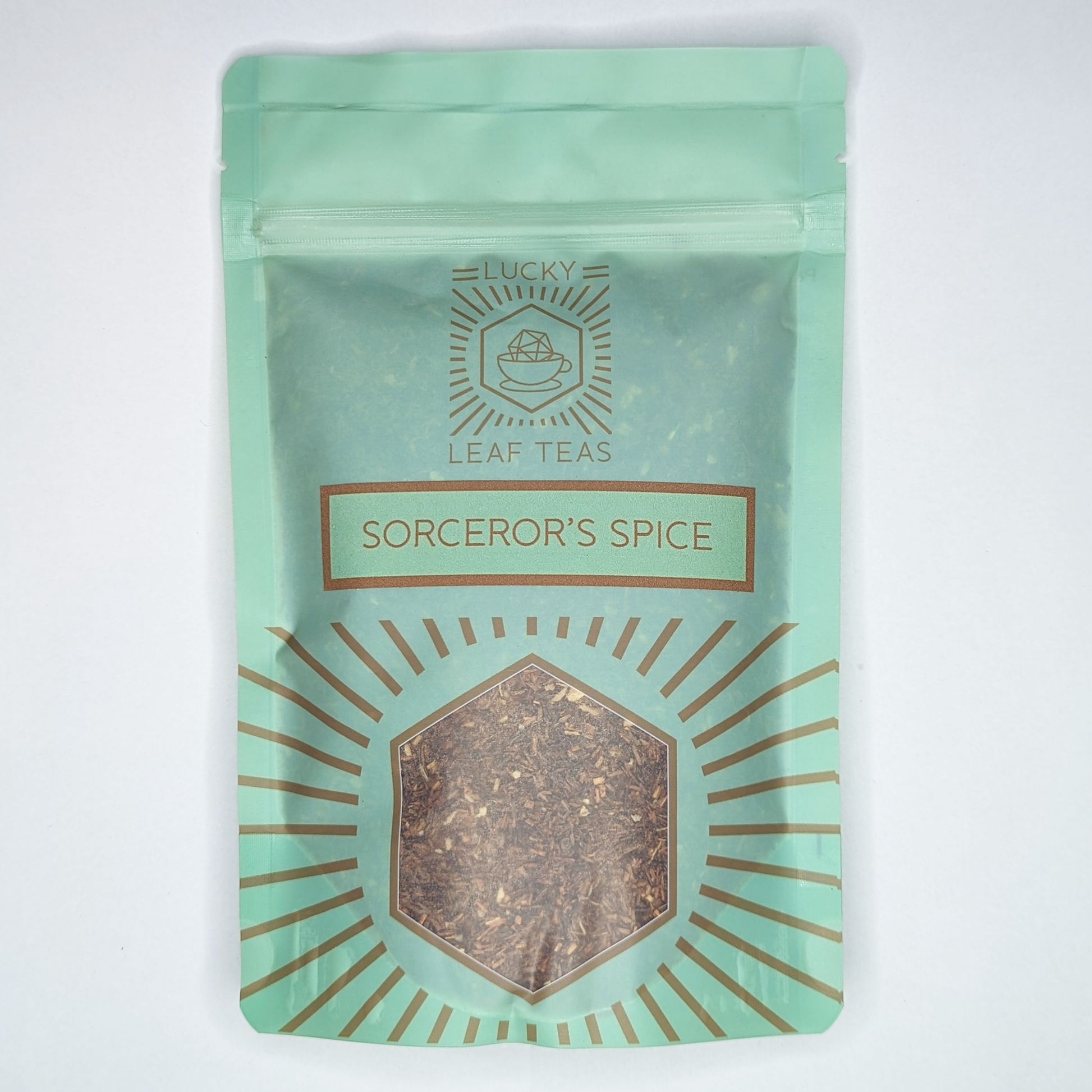 A mint-green package containing 100g of Sorcerer’s Spice: an infusion blend of Rooibos and spices. The name is stickered on the front, below the Lucky Leaf Teas Logo and above a hexagonal window displaying the tea.