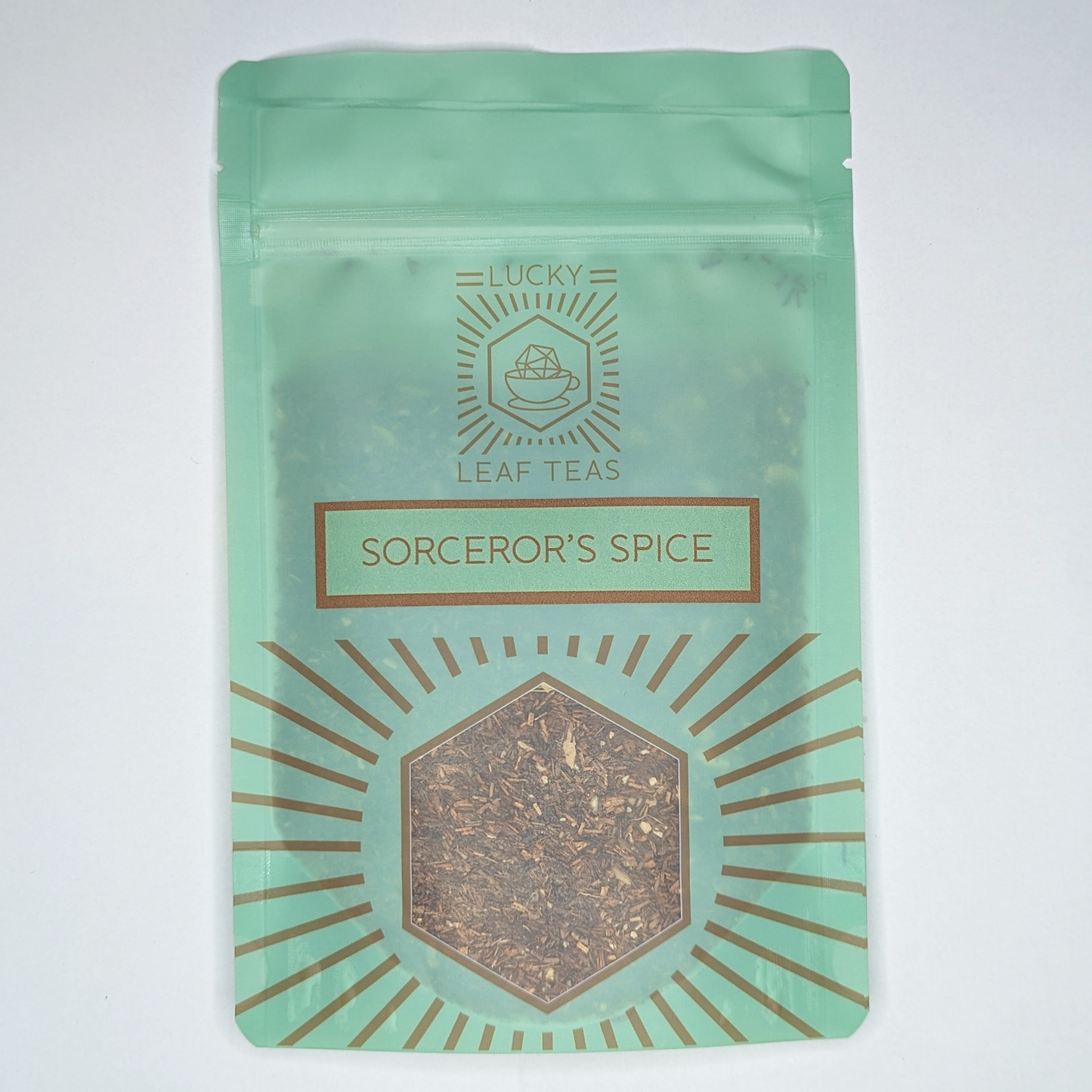 A mint-green package containing 50g of Sorcerer’s Spice: an infusion blend of Rooibos and spices. The name is stickered on the front, below the Lucky Leaf Teas Logo and above a hexagonal window displaying the tea.