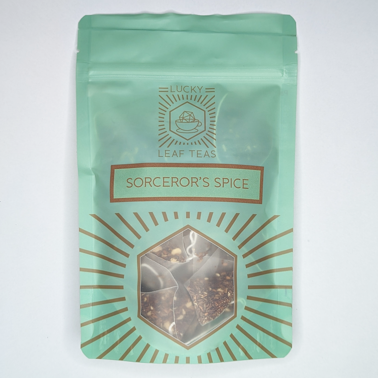 A mint-green package containing 15 Silky Pyramid Teabags of Sorcerer’s Spice: an infusion blend of Rooibos and spices. The name is stickered on the front, below the Lucky Leaf Teas Logo and above a hexagonal window displaying the tea.