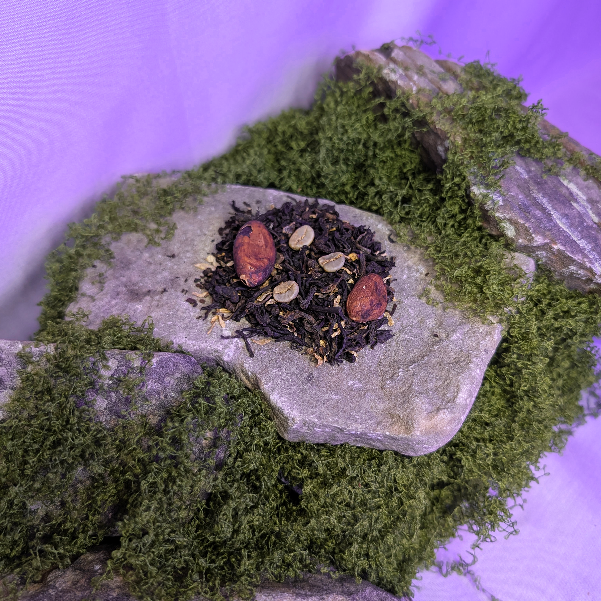 A small mound of tea leaves, with coffee and cocoa beans blended into it on top of a piece of grey slate. The slate itself is perched onto a heap of moss, and is surrounded by other pieces of slate.