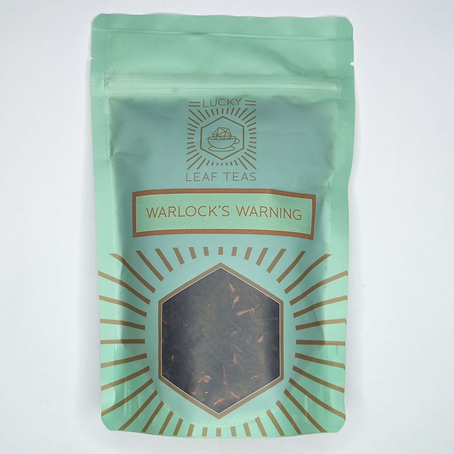 A mint-green package containing 100g of Warlock’s Warning: a black tea blend with visible red safflower petals. The name is stickered on the front, below the Lucky Leaf Teas Logo and above a hexagonal window displaying the tea.