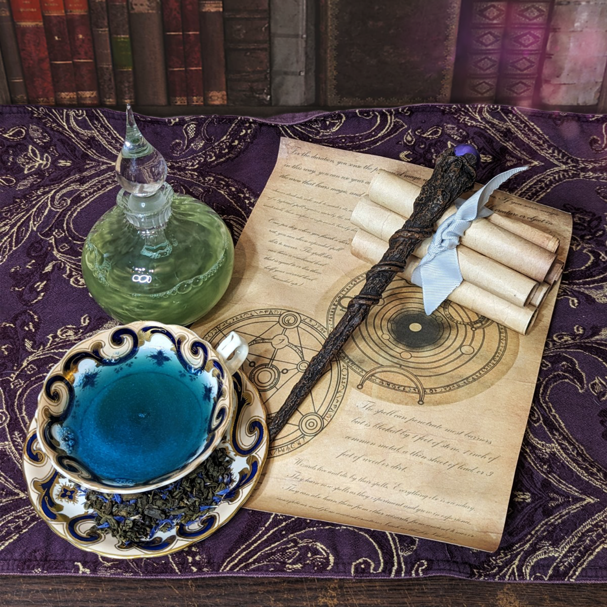 A glass potion bottle and white teacup on top of a spell scroll, next to a wand. The bottle is filled with shimmering green liquid, the cup filled with glittering blue liquid.