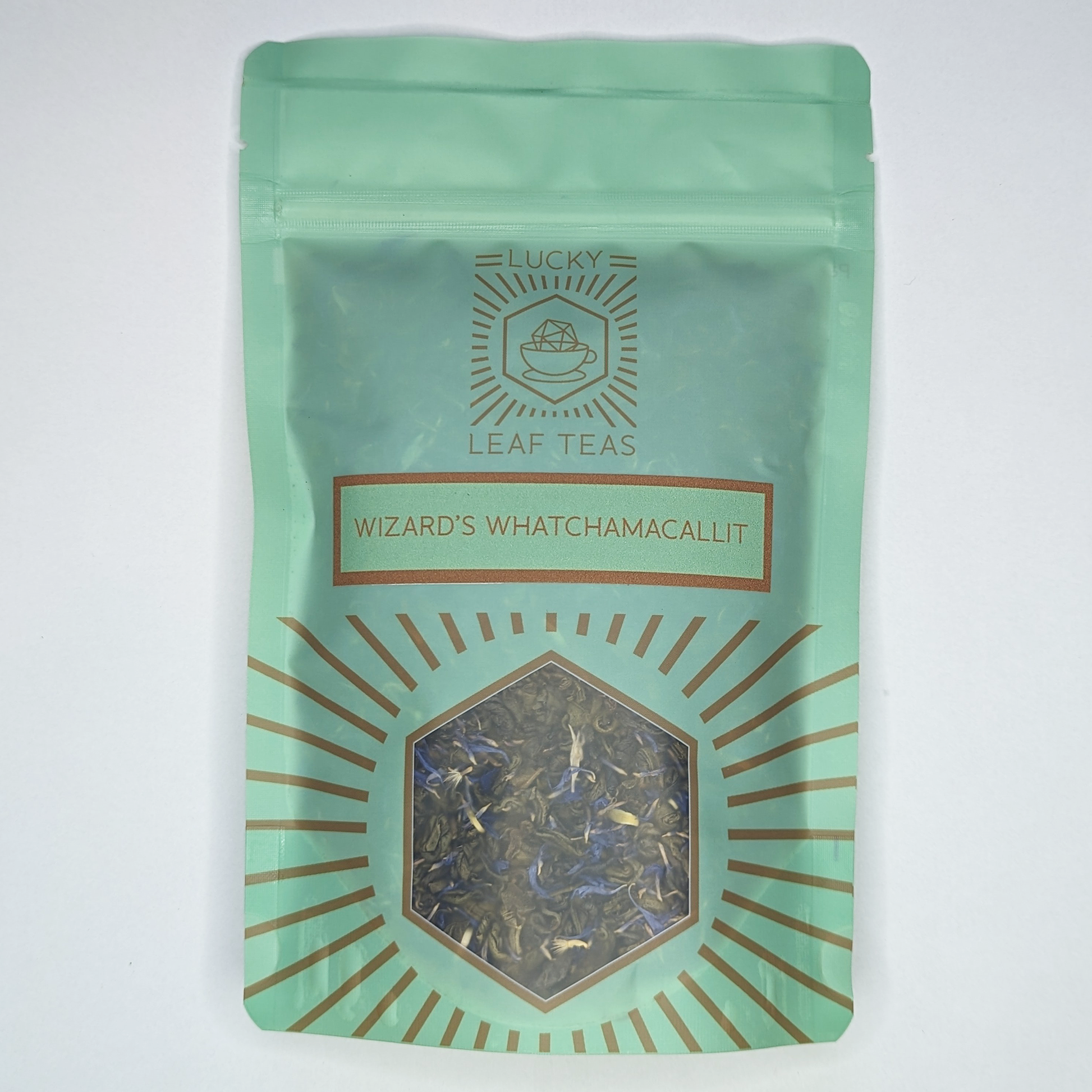 A mint-green package containing 100g of Wizard’s Whatchamacallit: a green tea blend with visible blue cornflower petals. The name is stickered on the front, below the Lucky Leaf Teas Logo and above a hexagonal window displaying the tea.