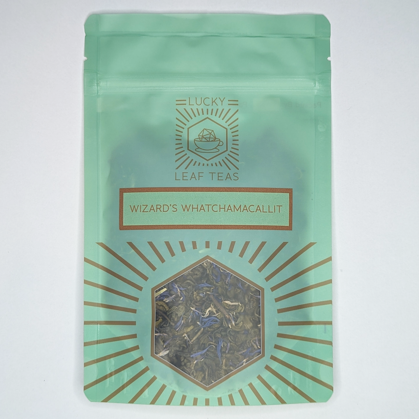 A mint-green package containing 50g of Wizard’s Whatchamacallit: a green tea blend with visible blue cornflower petals. The name is stickered on the front, below the Lucky Leaf Teas Logo and above a hexagonal window displaying the tea.